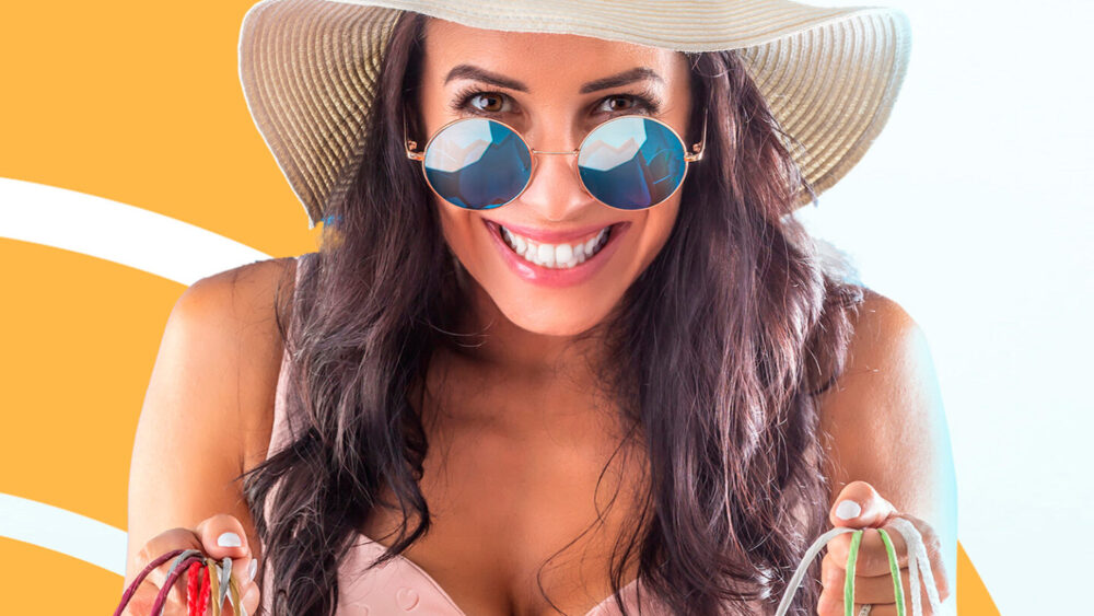 Mujer sonriente con sombrero de paja y gafas de sol, sosteniendo bolsas de compras porque se acerca la Semana Santa.